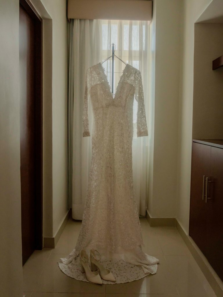 Lace wedding gown hanging by a window with bridal heels on the floor, representing the importance of professional bridal gown dry cleaning and preservation near Babylon